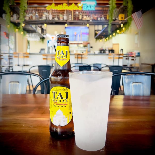 A bottle of Taj Mahal beer and a frosty glass on a wooden table in a bar setting.