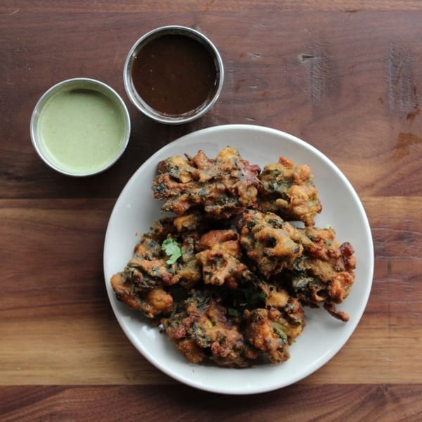 Crispy fried pakoras on a white plate with green and brown dipping sauces on a wooden table.