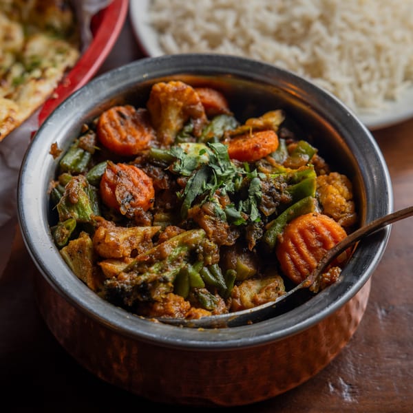 A copper bowl filled with Indian vegetable curry, garnished with cilantro, alongside rice and naan.