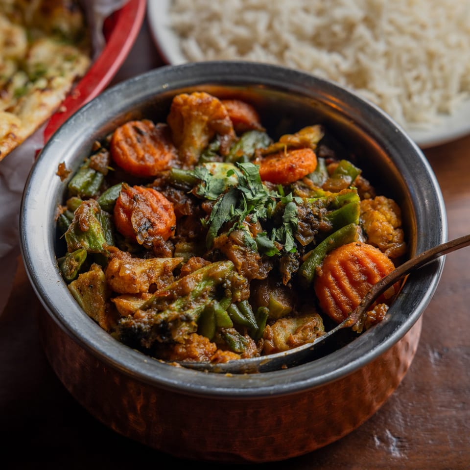 A copper bowl filled with Indian vegetable curry, garnished with cilantro, alongside rice and naan.