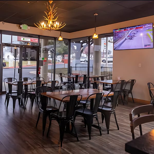 Restaurant interior with wooden tables, black chairs, large windows, and a TV showing a race.