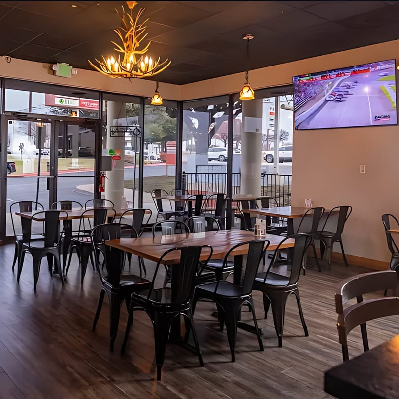 Restaurant interior with wooden tables, black chairs, large windows, and a TV showing a race.
