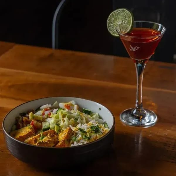 A colorful bowl of chicken, rice, and salad next to a red cocktail with a lime garnish.