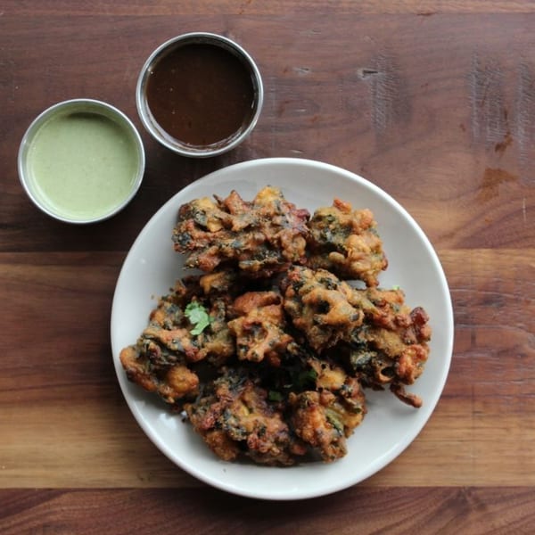 Crispy fried pakoras on a white plate with green and brown dipping sauces on a wooden table.