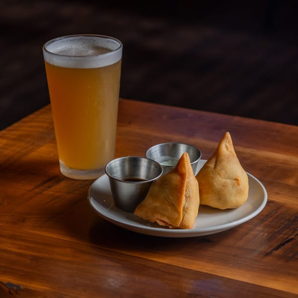 Two golden-brown samosas with dipping sauces on a white plate next to a glass of beer.