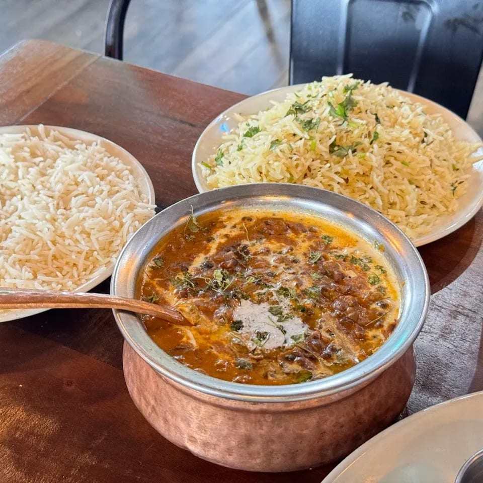 A copper pot of creamy dal makhani with two plates of rice on a wooden table.