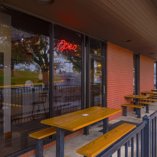 Outdoor patio with wooden picnic tables and benches in front of a brick building with an "Open" sign.