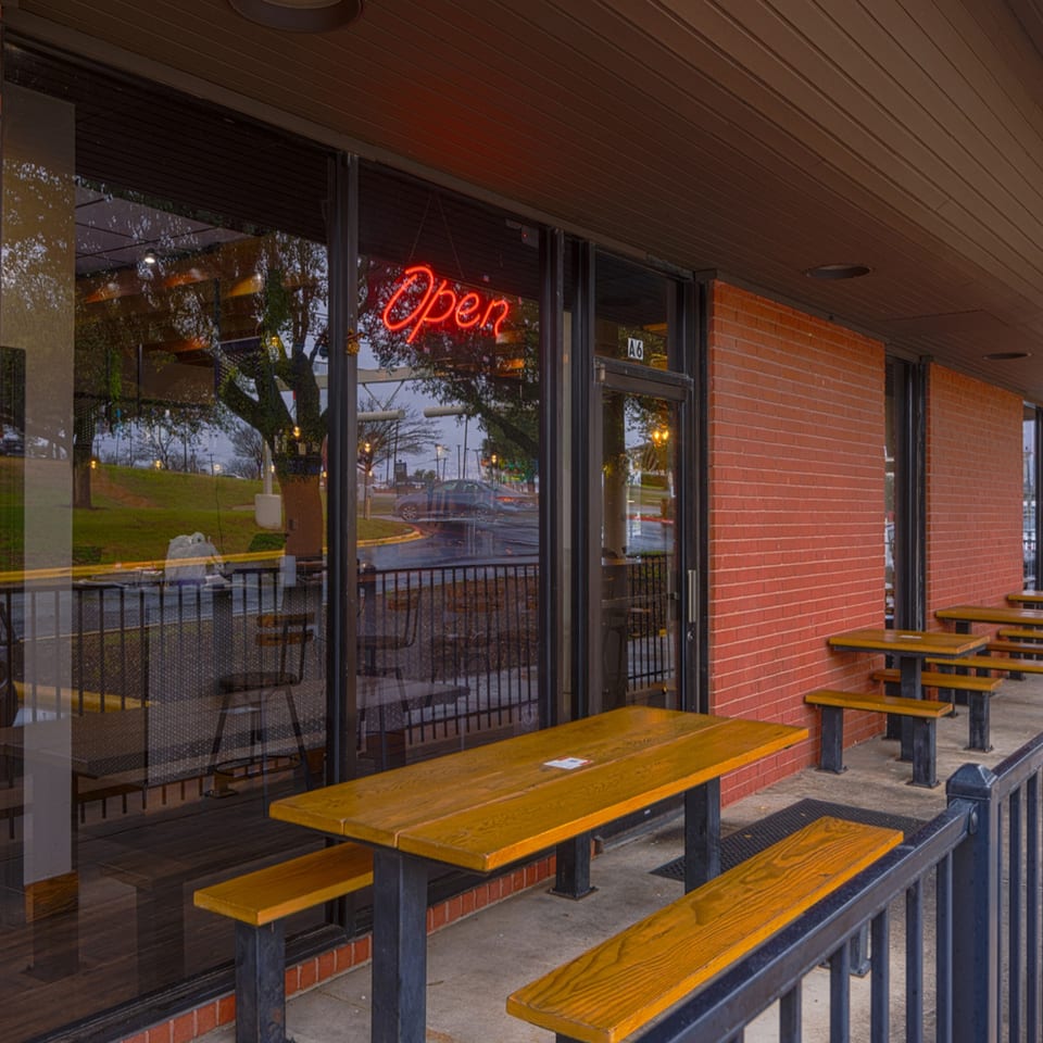 Outdoor patio with wooden picnic tables and benches in front of a brick building with an "Open" sign.