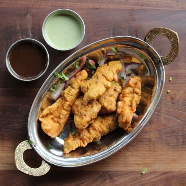 Golden fried pakora with red onions and cilantro in a silver dish, with two dipping sauces.
