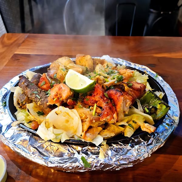 Sizzling platter of mixed grilled meats, vegetables, and a lime wedge on a foil-lined plate.