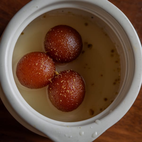Three golden-brown gulab jamuns, sprinkled with nuts, soaking in a sweet syrup in a white bowl.