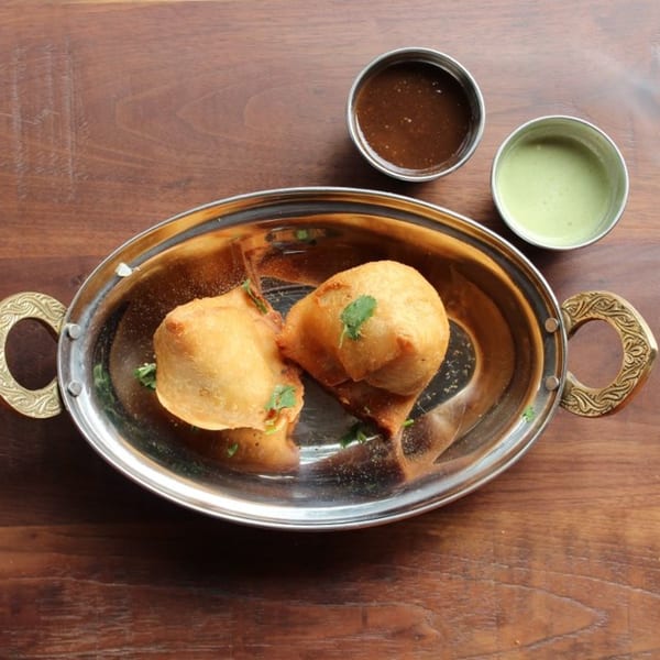 Two golden-brown samosas garnished with cilantro in a silver serving dish, with two small bowls of dipping sauces.