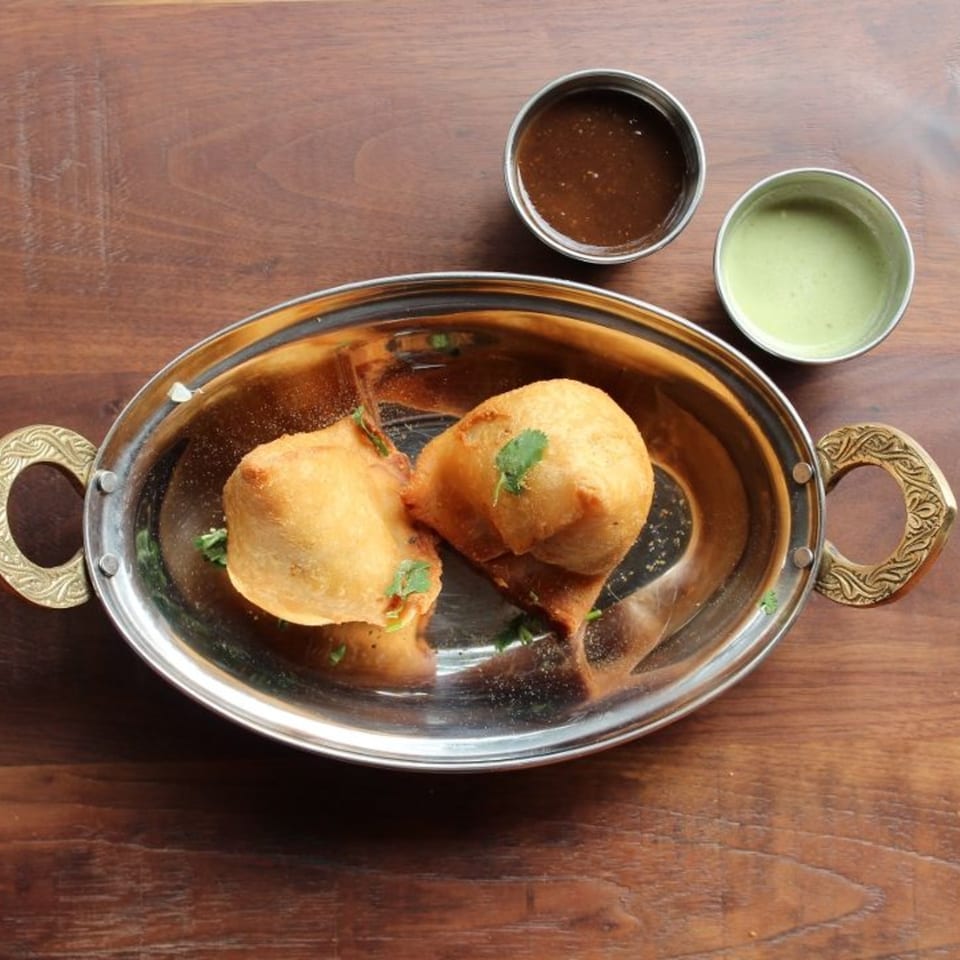 Two golden-brown samosas garnished with cilantro in a silver serving dish, with two small bowls of dipping sauces.