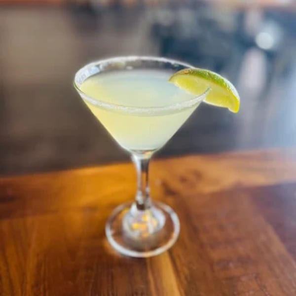 A margarita in a martini glass with a salted rim and a lime wedge on a wooden table.