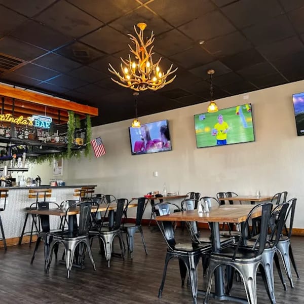 Restaurant interior with a bar, wooden tables, metal chairs, TVs, and an antler chandelier.